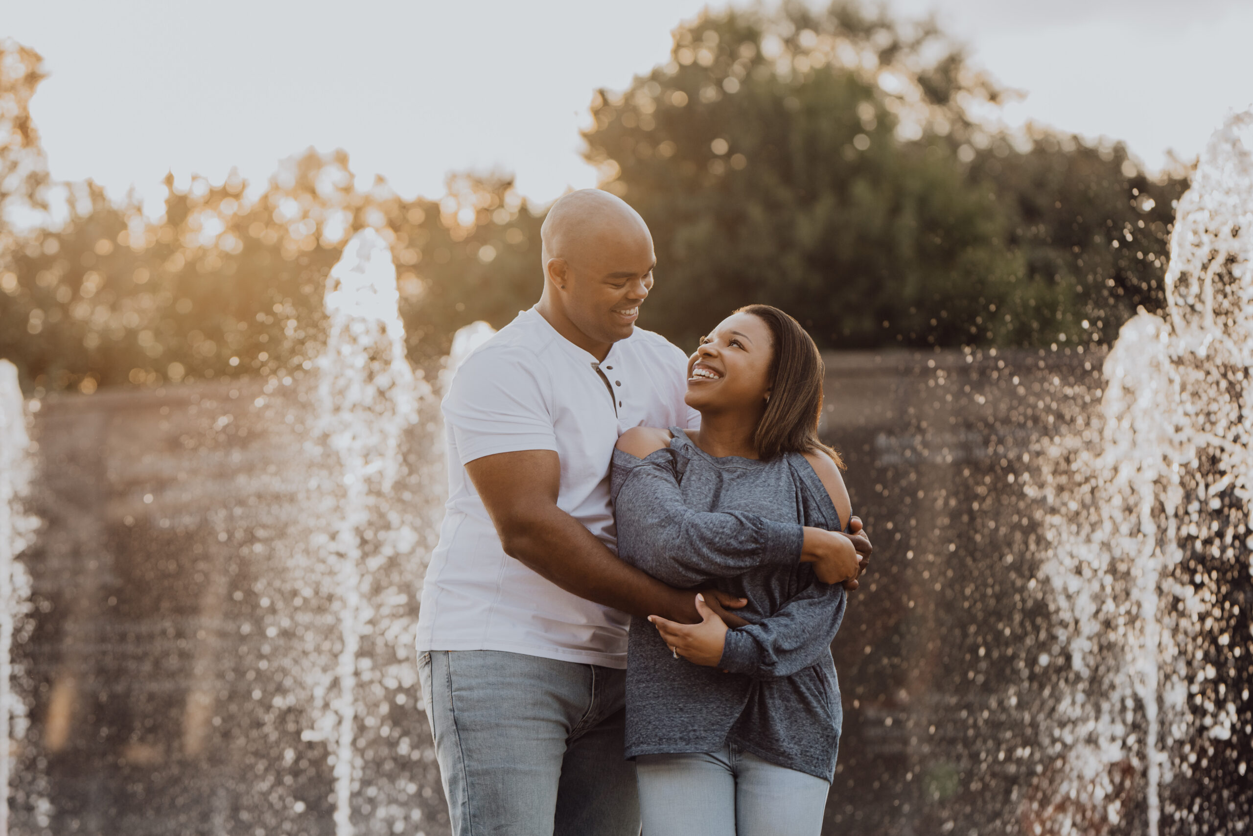 a man and woman holding each other and smiling and looking at each other with a fountain and sunlight behind them at their engagement session in Nashville