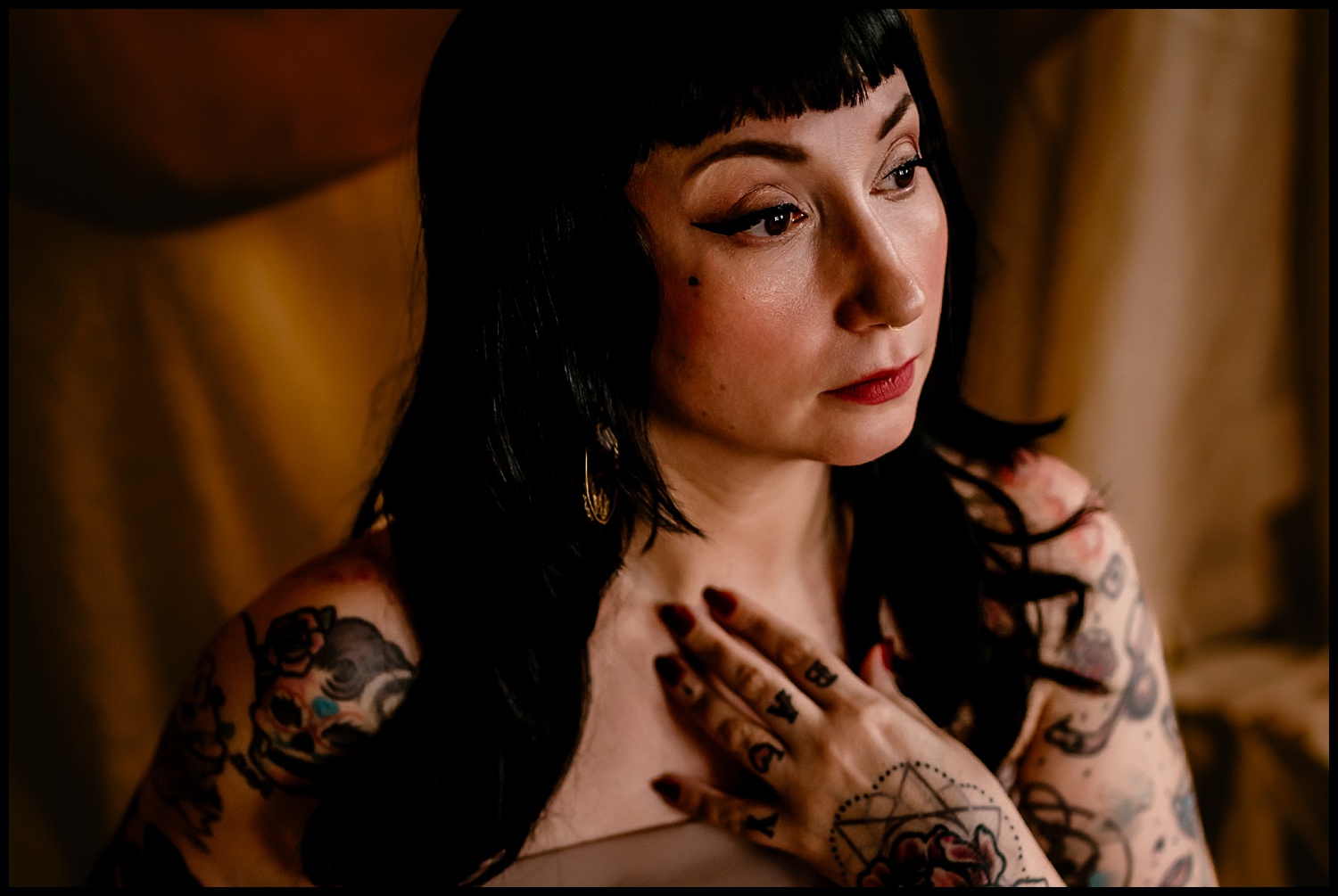 The person with black hair styled in a wet look gazes thoughtfully to the side, tattoos adorning their arms and a ring tattoo on their finger. Their hand rests on their chest against a softly blurred background imbued with warm, classical tones.