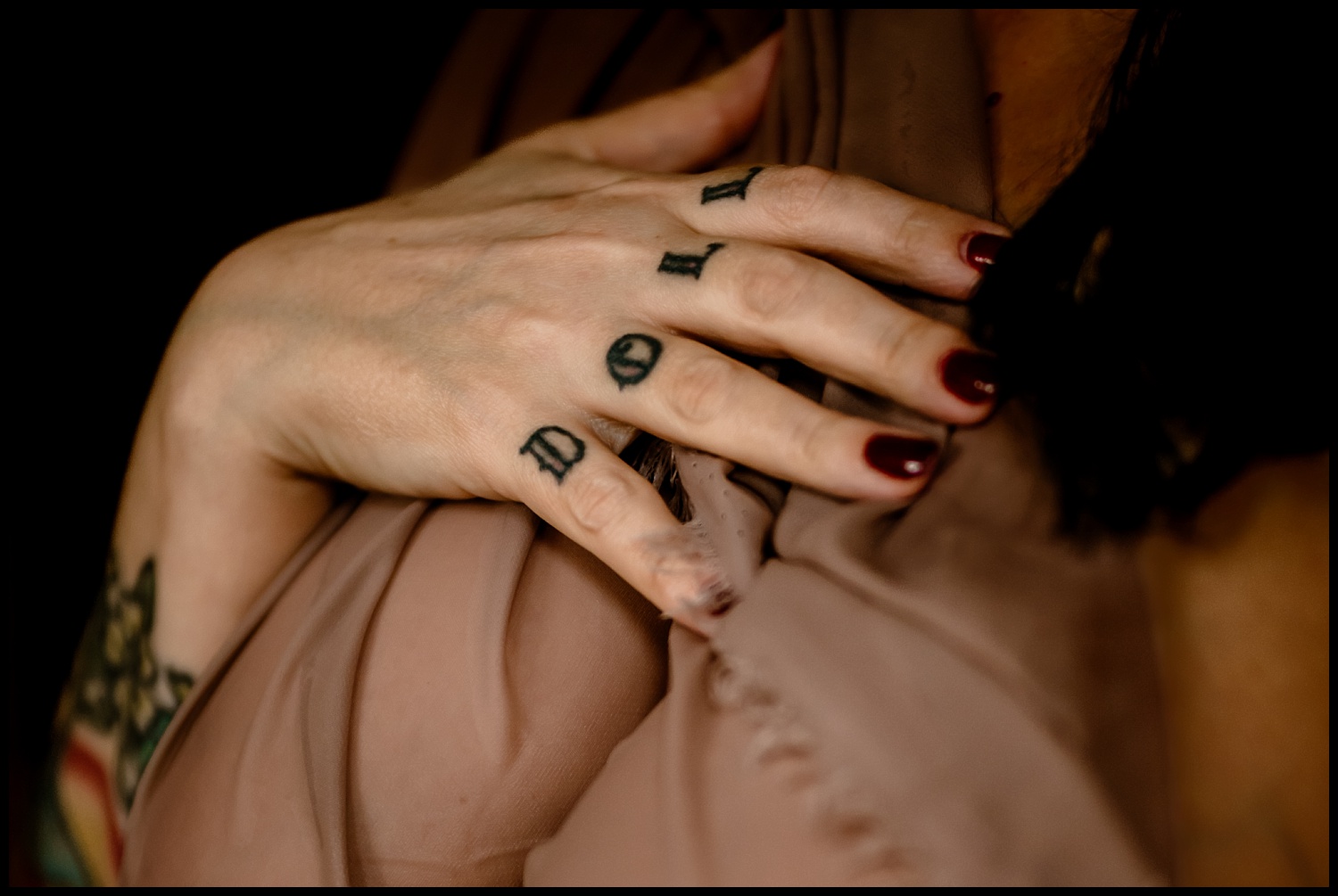 Close-up of a hand with DOLL tattooed on the fingers, resting elegantly on sheer brown fabric. The persons nails boast a dark red polish, complementing a vibrant wrist tattoo. This fashion-forward moment is set against a dark, blurred backdrop, adding a touch of classical allure.