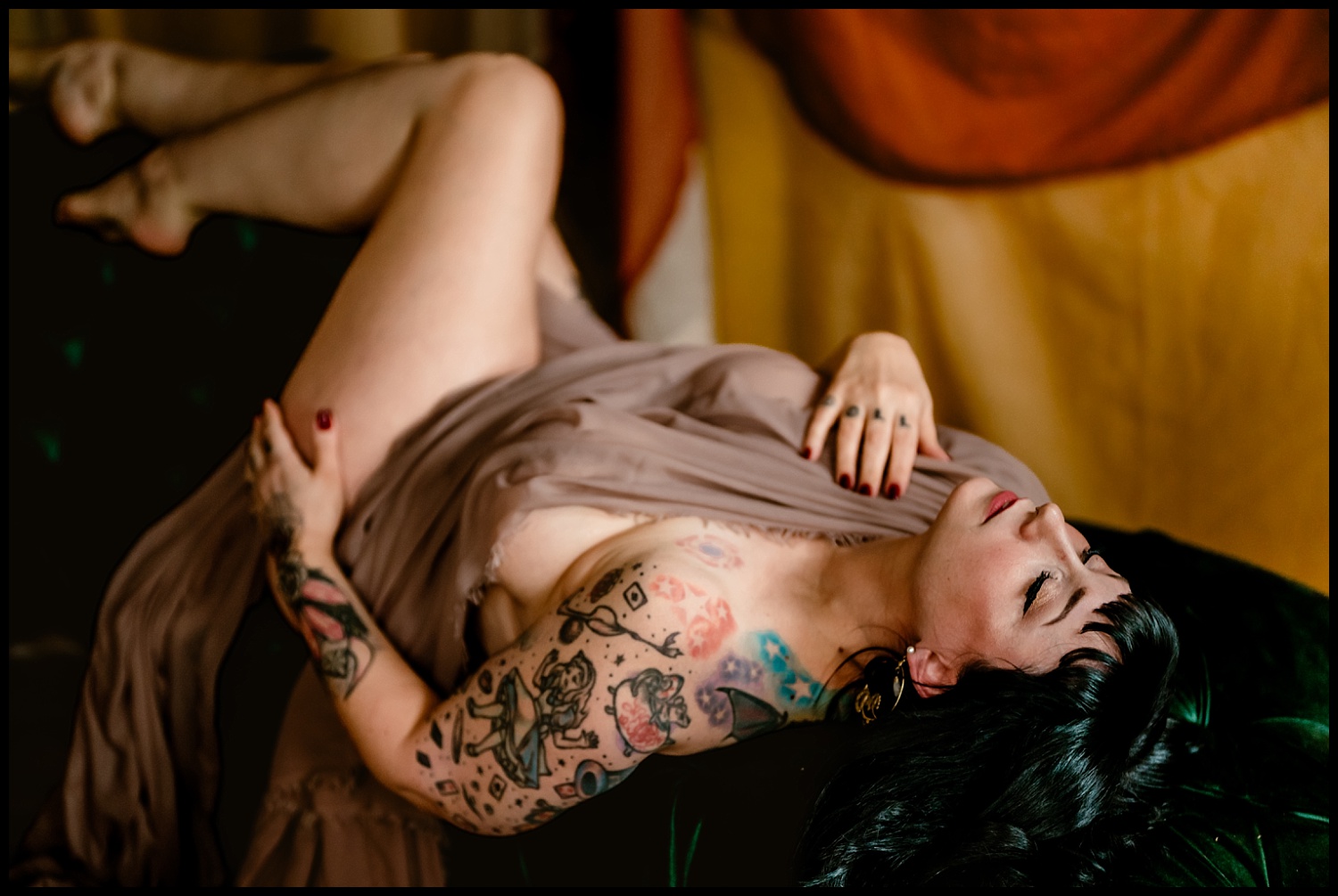 A person with tattoos reclines on a dark green sofa, their dark hair styled in a wet look. Draped in sheer beige fabric, they have a relaxed expression with one leg bent up. The background evokes a classical feel with its warm, muted colors.