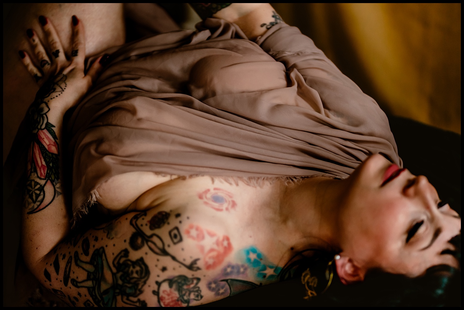 A person with tattoos lies on their back, partially covered with a beige fabric. The colorful tattoos are detailed and evoke a classical feel. The lighting is warm, creating a soft and intimate atmosphere reminiscent of a timeless painting.