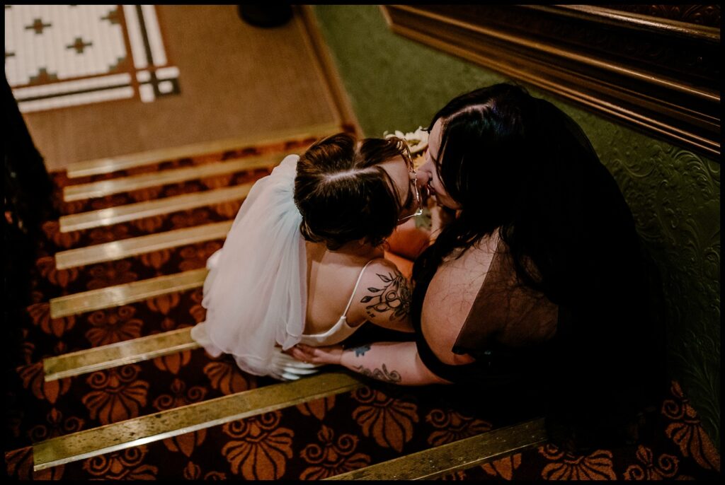 A bride in a white dress and veil sits on the patterned carpeted stairs, facing her partner in a dark outfit. They share a romantic wedding portrait moment, gazing into each others eyes, surrounded by the textured wall that adds to the scenes intimacy.