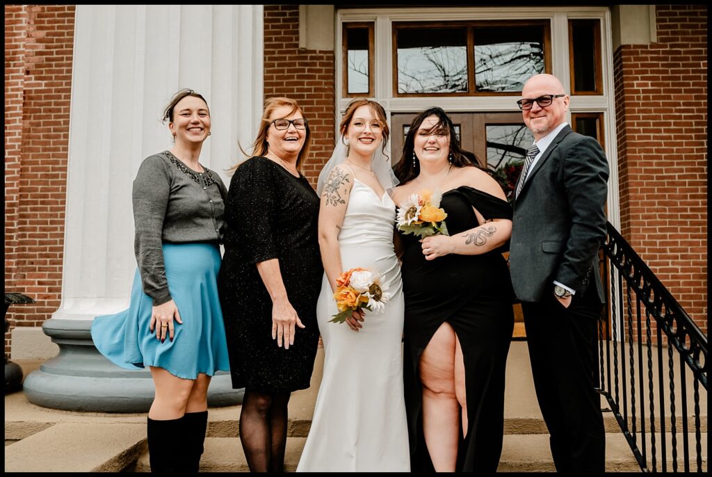 A group of five people stands on steps in front of a brick building with large windows. They are dressed formally, with two holding bouquets. The scene captures the joy of courthouse wedding portraits, suggesting a celebration filled with laughter and romance.