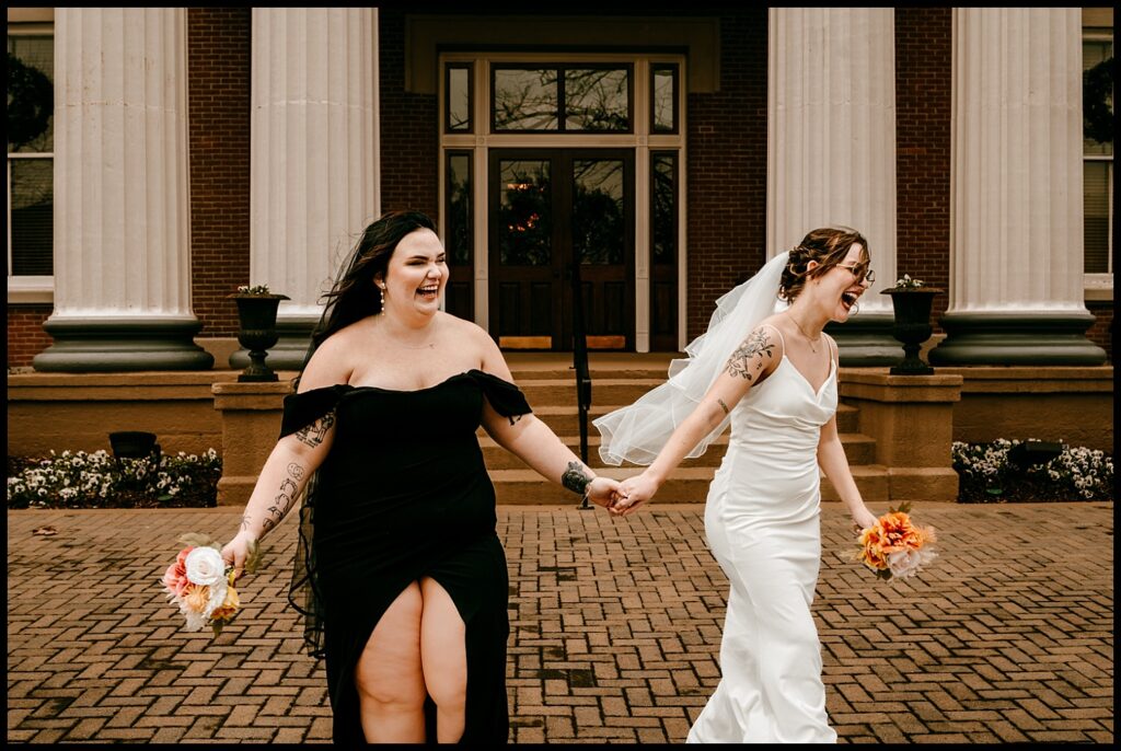 Two brides, in a romantic portrait outside a courthouse with columns, hold hands and laugh. One is in a black dress, the other in a white gown and veil. Their bouquets complement their joy-filled expressions, perfectly capturing the essence of laughter-filled celebration.