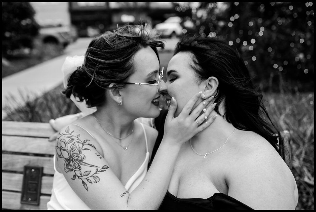 Two people sitting on a bench are about to kiss, their eyes meeting with the warmth of a courthouse wedding. One has a floral tattoo, adding charm to this romantic portrait in an intimate black-and-white outdoor setting. Joy and love fill the air as they share this tender moment.