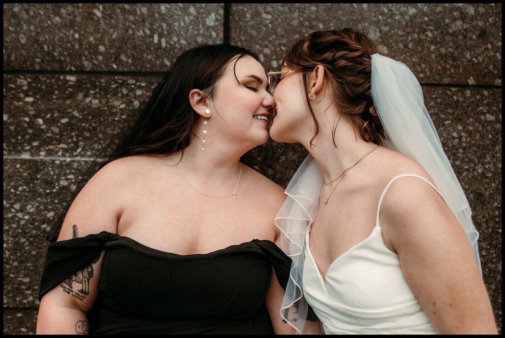 Two women, one in a black dress and the other in a white dress with a veil, lean in for a kiss against a textured stone wall. Their joyful expressions capture the essence of laughter-filled portraits, showcasing an intimate moment of affection and celebration.