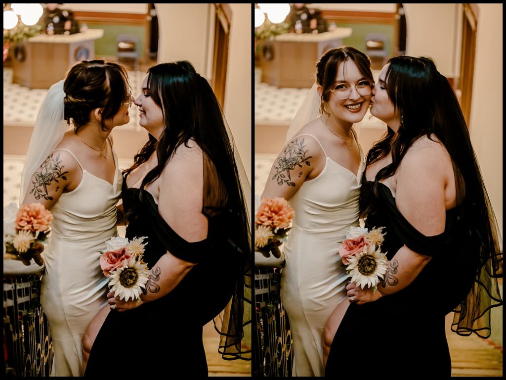 A lesbian couple in wedding attire stand closely on a staircase, capturing the charm of romantic portraits. The one in white, with a veil and bouquet, smiles as the one in black kisses her cheek and holds flowers. Both have tattoos, with the elegant setting featuring a patterned floor beneath them.