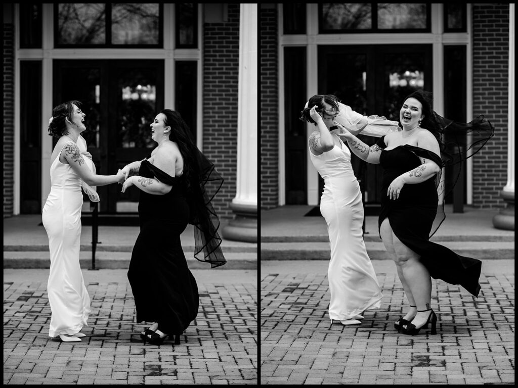 A lesbian couple in wedding dresses joyfully dance together on a brick path, embodying laughter-filled wedding portraits. One wears a white dress; the other, a black dress with a flowing veil. They laugh and spin, expressing pure happiness and celebration.