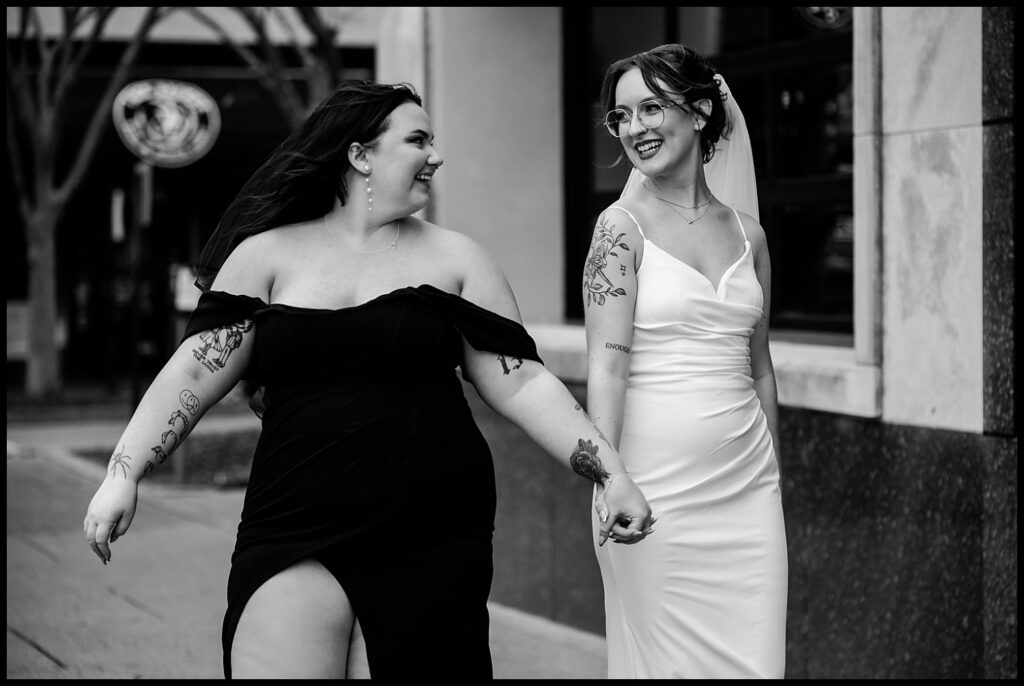Two brides hold hands, walking outdoors in a laughter-filled scene. One wears a wedding dress with veil, the other an off-the-shoulder black dress. Tattooed and bespectacled, they smile warmly at each other, exuding happiness in this romantic black-and-white image.