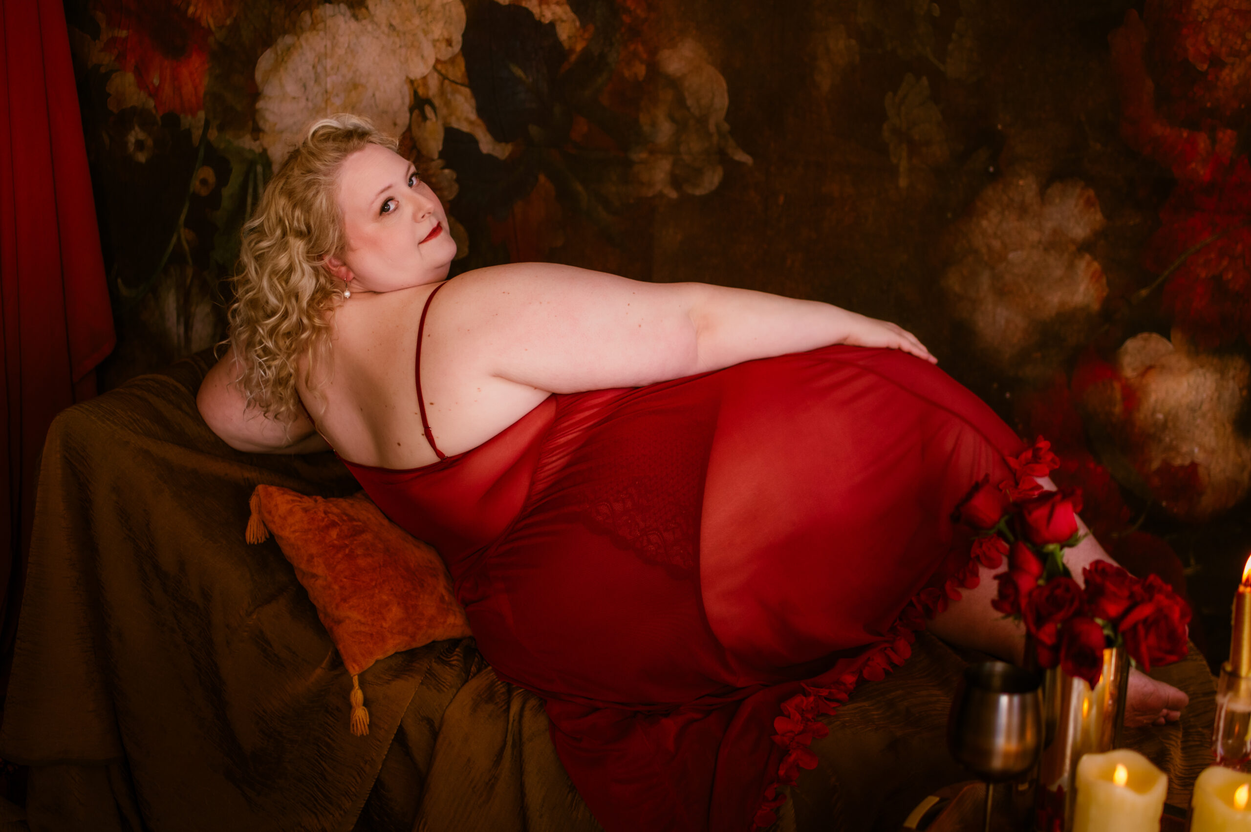 A woman with curly blonde hair lounges elegantly on a couch, wearing a flowing red gown. The floral backdrop evokes a Renaissance charm. The setting is warm and softly lit, creating a luxurious and intimate boudoir atmosphere, like something from a dream.