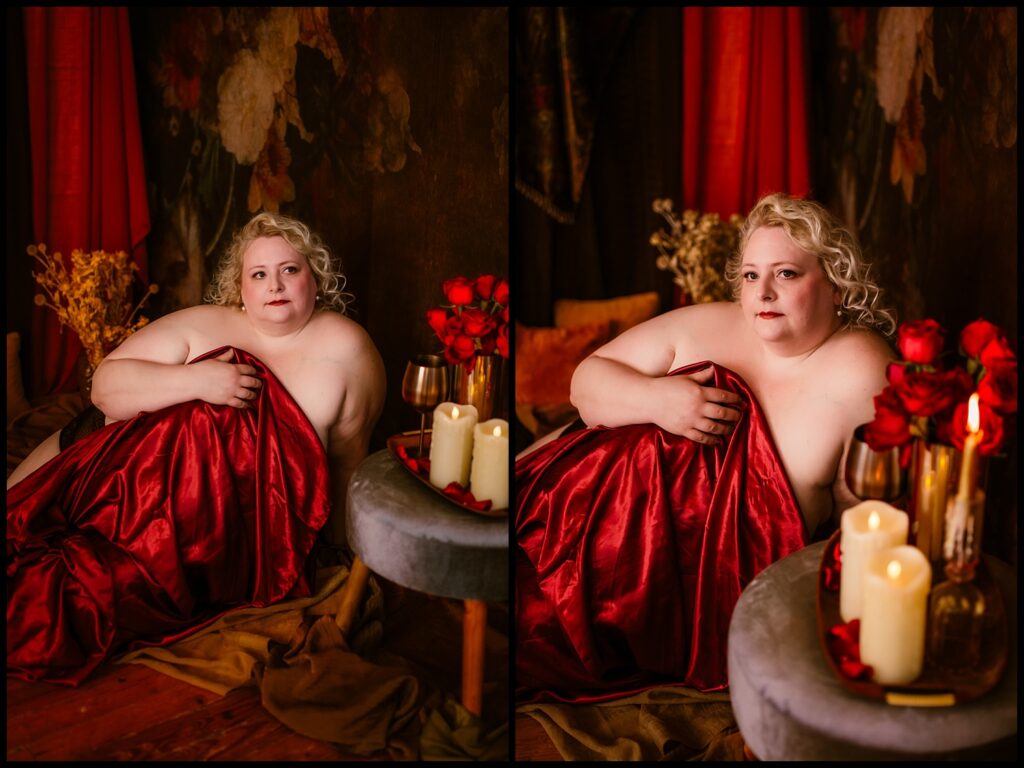 A person with curly blonde hair sits on a wooden floor, wrapped in a red satin sheet. The scene is reminiscent of a Renaissance dream, surrounded by flickering candles, a wine glass, and red flowers against dark floral decor and rich red curtains.