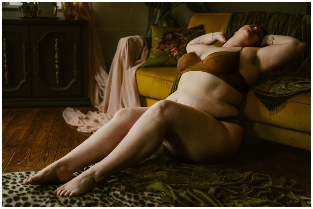 A person in brown lingerie reclines against a yellow sofa, eyes closed and arms raised, with soft light highlighting their body—an intimate moment captured during a boudoir session. Fabric and a green blanket are draped nearby on a wooden floor.
