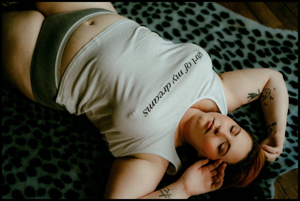 A woman with tattoos is lying on her back on a leopard-print blanket, wearing a white tank top that reads girl of my dreams and grey underwear. This boudoir session captures her relaxed state, eyes closed and one hand near her face.