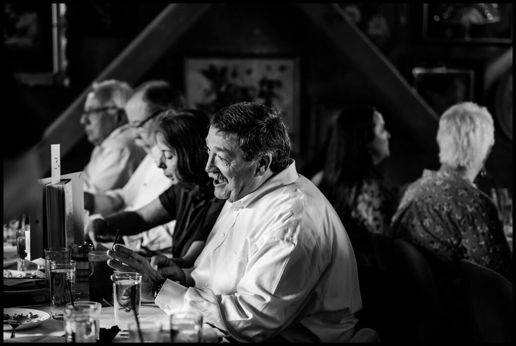 A group of people, including Jess and Chris, sit at a long table in a restaurant, engaged in conversation and laughter. The black and white scene focuses on a smiling man in a white shirt gesturing with his hands.