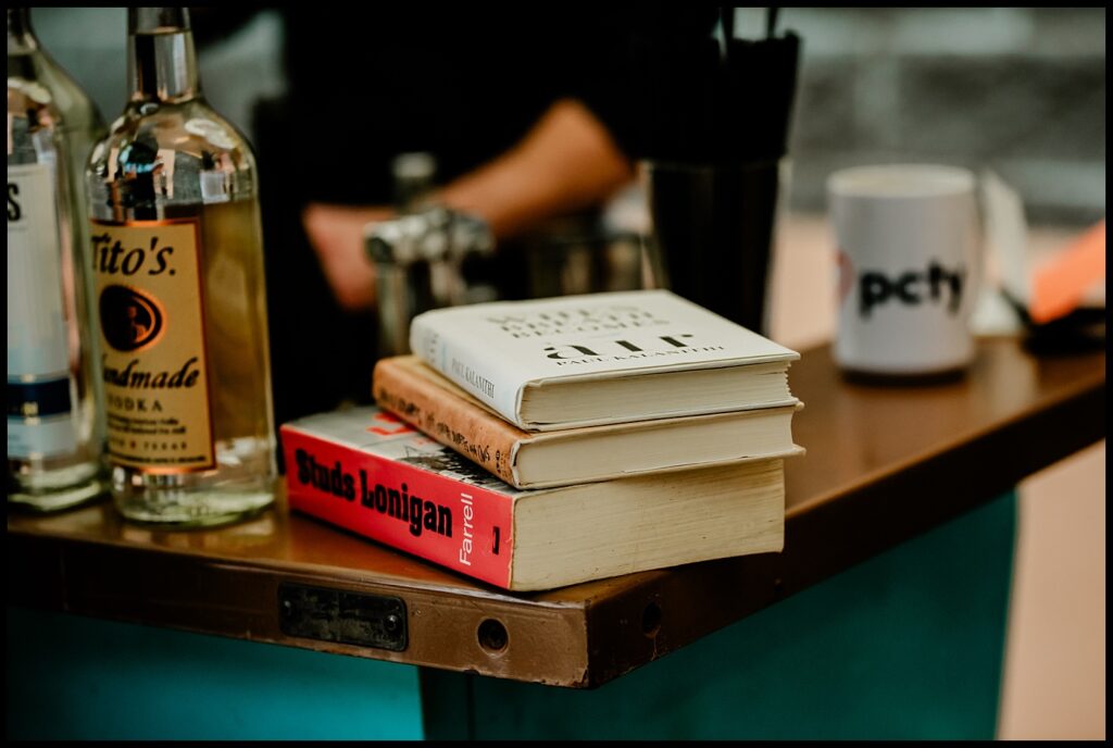 A stack of three books sits on a bar counter next to bottles of Tito’s Handmade Vodka, a coffee mug, and bar utensils; Jess and Chris stand blurred in the background.