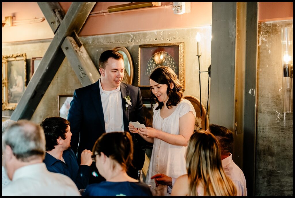 Jess and Chris stand together, he in a suit and she in white, as she reads a card, both smiling amid warmly lit rustic decor, framed art, and their seated guests.