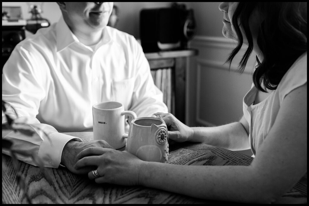 A black-and-white photo of Jess and Chris sitting at a table, holding hands and smiling at each other. Both have mugs in front of them. The woman wears a ring, and the scene feels warm and intimate.