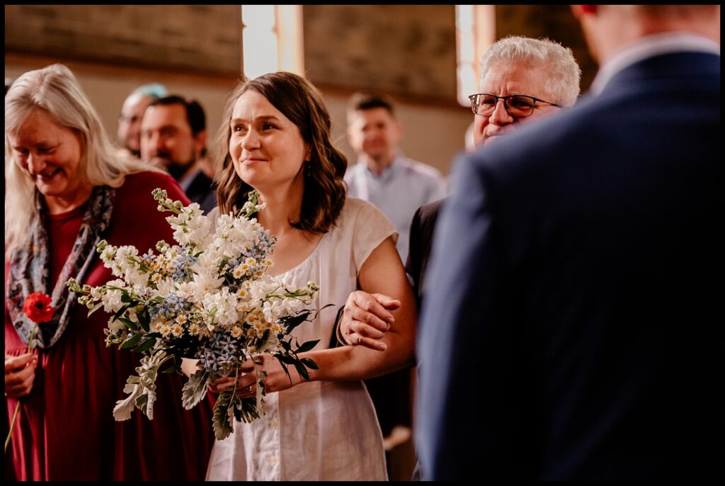 Jess and Chris’s special day unfolds as a bride holding a bouquet of white and blue flowers walks down the aisle, smiling, arm-in-arm with an older man. Guests watch and smile in the background inside a warmly lit venue.