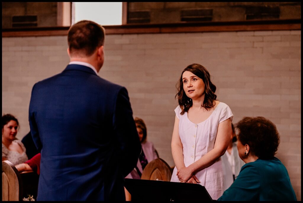 Jess, wearing a white dress, stands and speaks to Chris, who is in a suit with his back to the camera. Several people are seated around them in a room with brick walls, giving the scene a formal or ceremonial feel.
