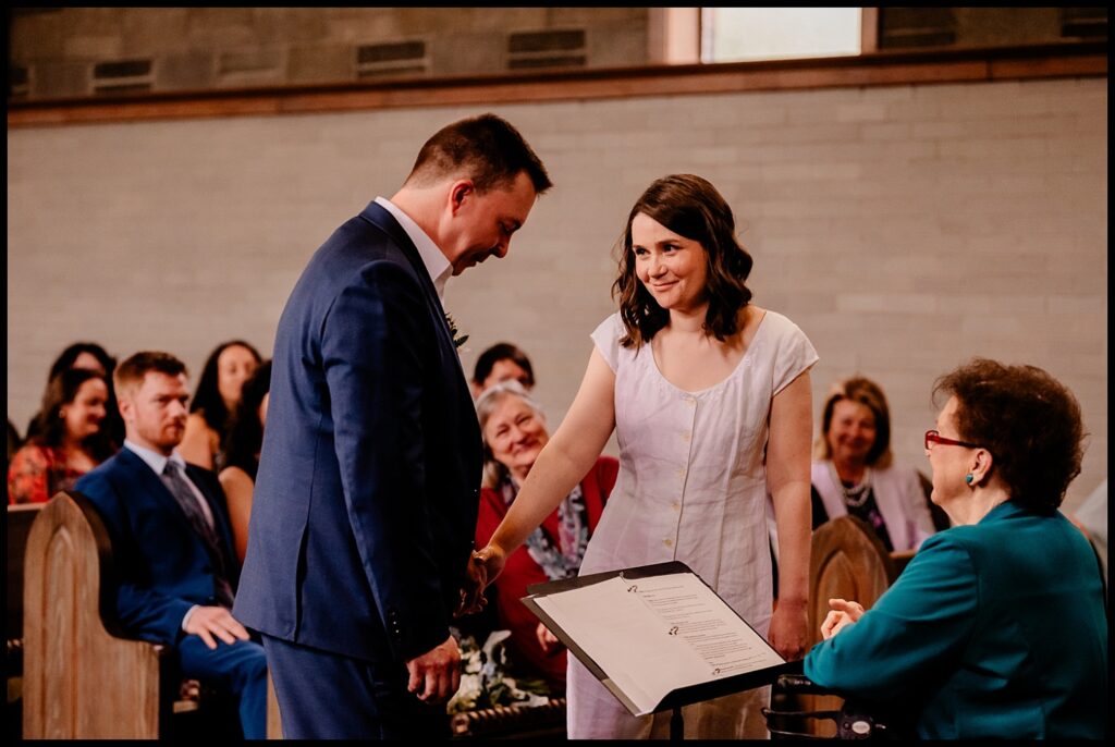 Jess and Chris stand at the altar, holding hands and smiling at each other during their wedding ceremony, while a woman in teal officiates and guests watch from the pews in the background.