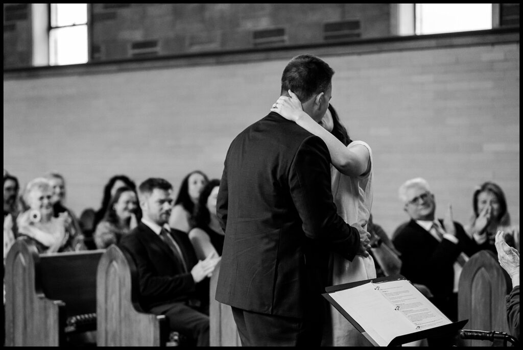Jess and Chris share a kiss at the front of a church as guests seated in pews smile and applaud. The scene is in black and white, with sheet music visible in the foreground.
