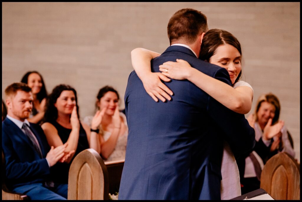 Jess and Chris embrace warmly in a church pew as people seated behind them watch and applaud, some smiling. The scene is joyful and intimate, suggesting a special or celebratory moment.