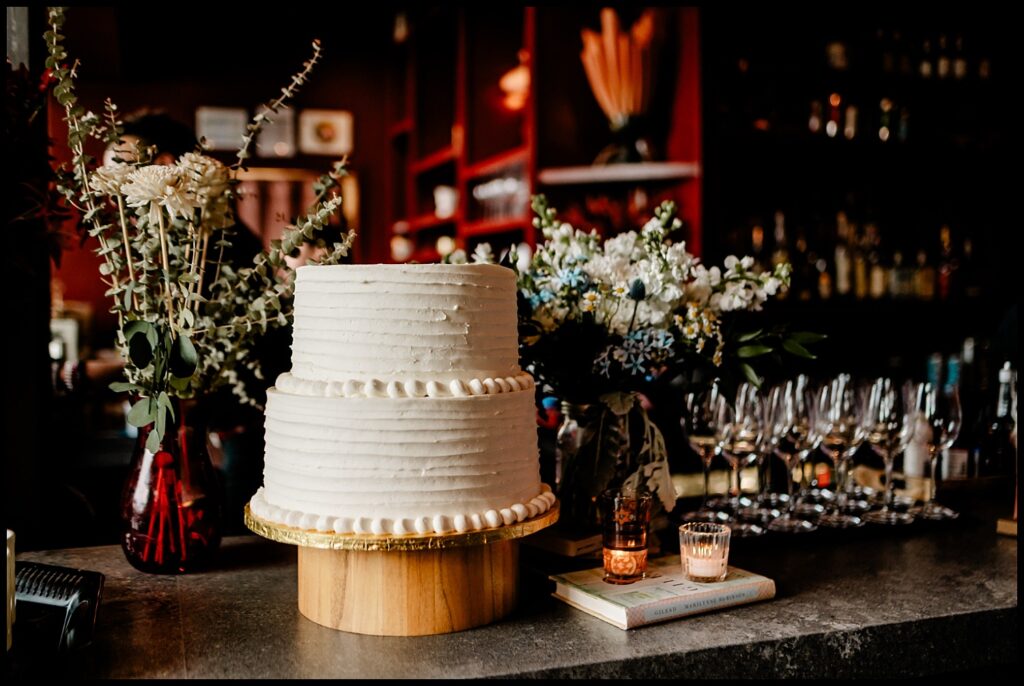 A two-tiered white frosted cake sits on a wooden stand on a bar counter, surrounded by flower arrangements, wine glasses, candles, and books in a warmly lit, elegant setting—perfect for Jess and Chris’s celebration.