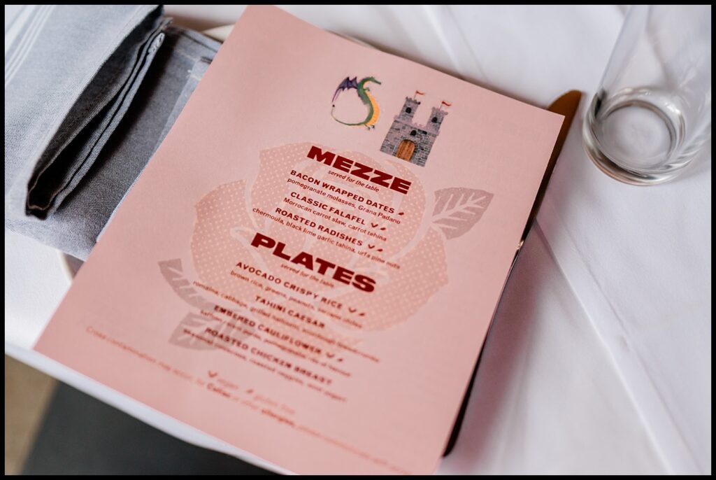 A pink restaurant menu for Jess and Chris, with a rose graphic and small dragon illustration, lists “Mezze” items like bacon-wrapped dates and falafel, plus “Plates” including avocado crispy rice and grilled cauliflower.