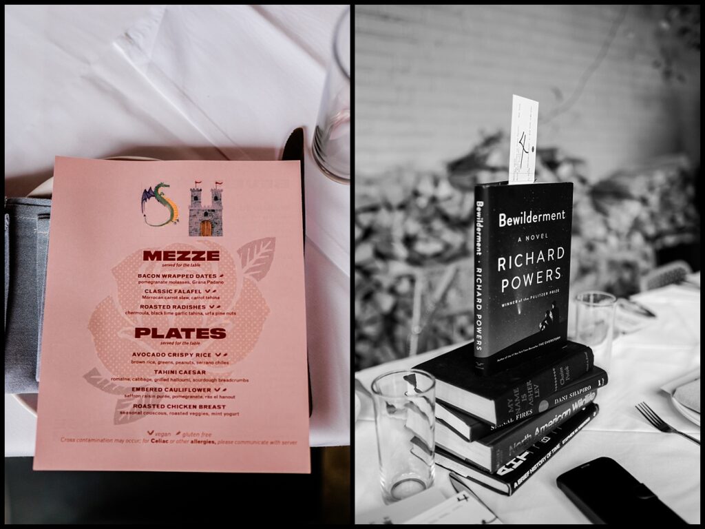 A close-up of a restaurant table setting: on the left, a pink menu listing mezze and plate options; on the right, a stack of books—Jess and Chris’s favorites—topped with Bewilderment by Richard Powers, used as table decor.
