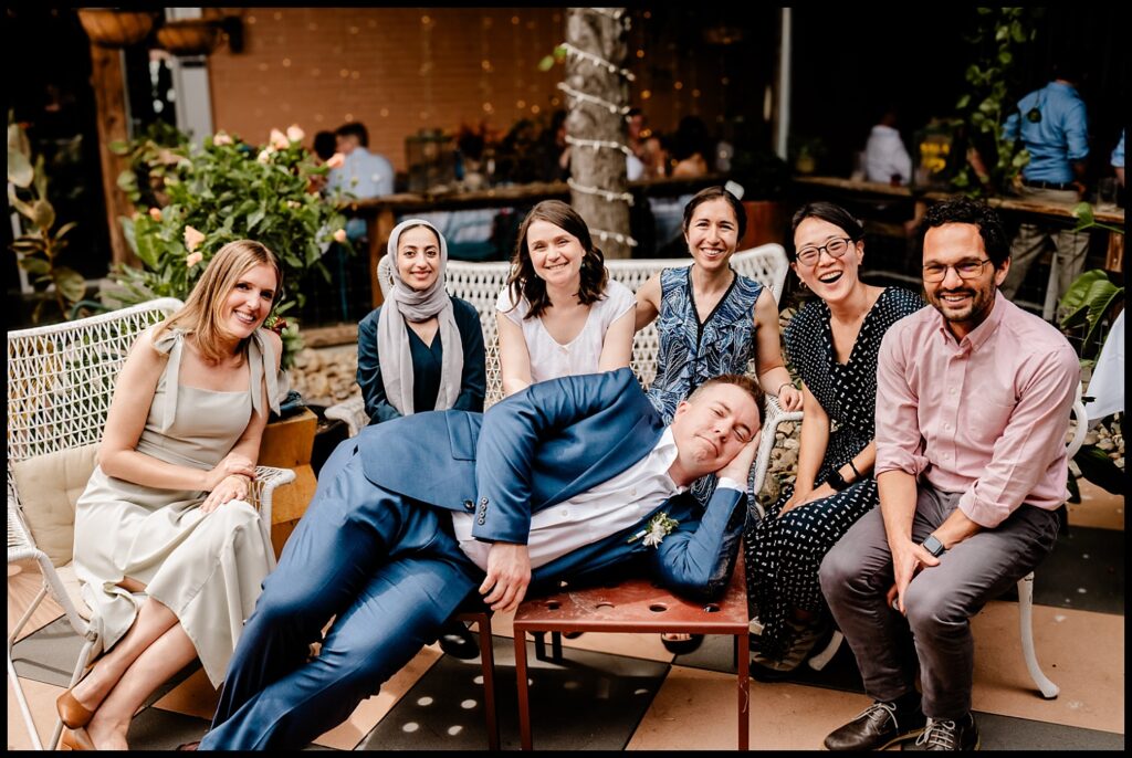 Jess and Chris are among seven people smiling for a group photo outdoors. Six sit on benches, while one man in a blue suit playfully lies across a table, resting his head on his hand. The setting is casual and cheerful.