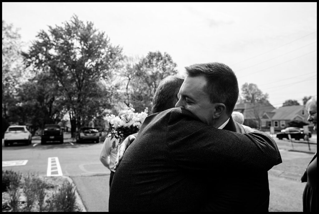 Jess and Chris embrace in a heartfelt hug outdoors on a street, one holding a small bouquet of flowers. The background shows parked cars, trees, and houses under a partly cloudy sky. The image is in black and white.