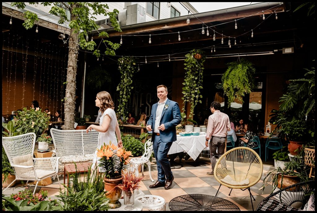 Jess and Chris stand in a lush, outdoor patio area decorated with plants, white chairs, and string lights—a man in a blue suit and a woman in a white dress—while two others mingle near a table in the background.