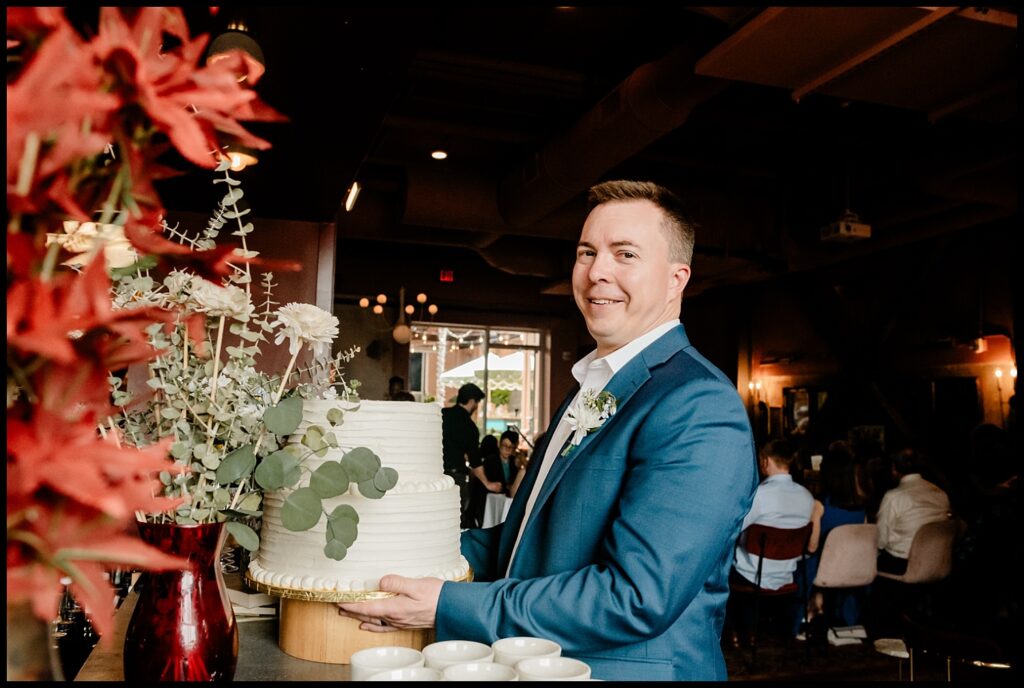 A smiling man in a blue suit holds a white tiered cake near a large bouquet of flowers inside a dimly lit room, celebrating Jess and Chris as people are seated in the background.