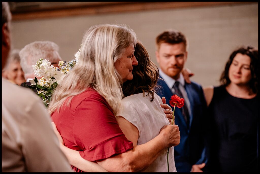 Jess and Chris share an emotional hug, surrounded by supportive people. Among them, a man in a suit holds a red flower, adding to the heartfelt atmosphere.