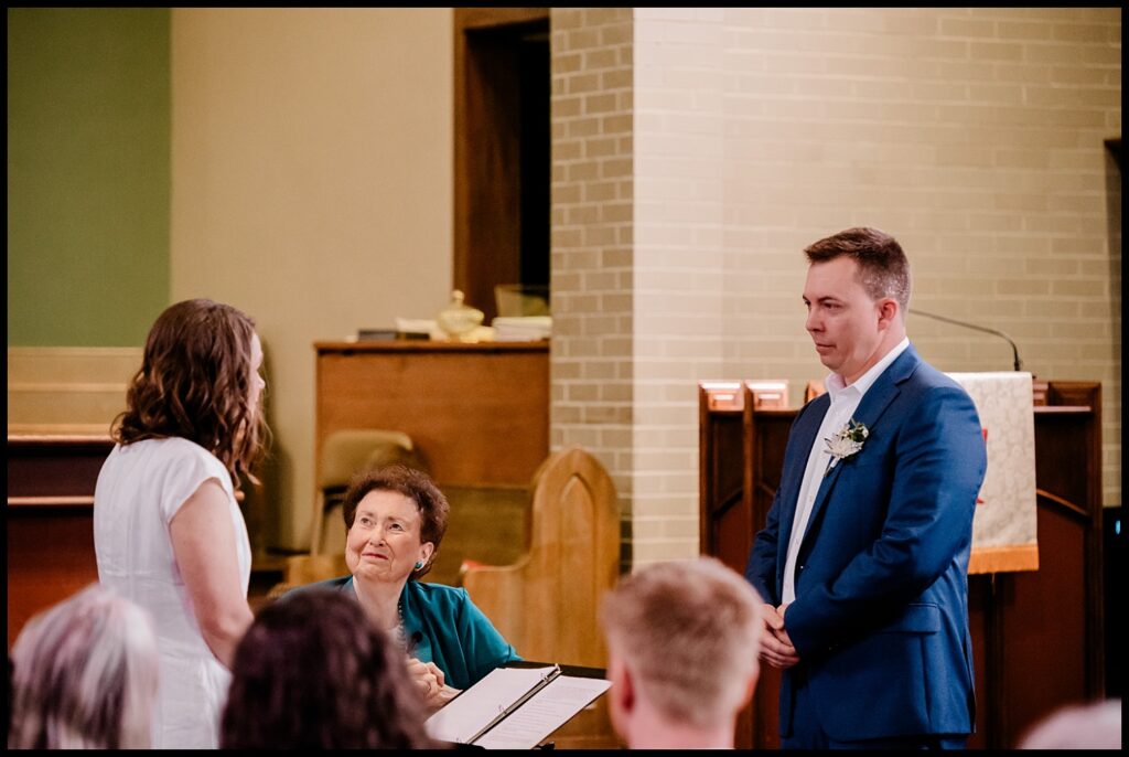 Jess and Chris stand facing each other during their wedding ceremony, with an officiant seated between them, holding a book. The indoor setting features wooden pews and a brick wall in the background.