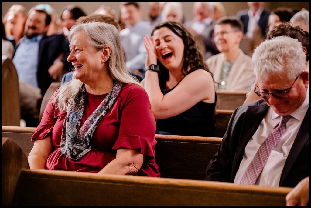 People seated in church pews, including Jess and Chris, are smiling and laughing. A woman in a red dress is in the foreground, while others behind her enjoy the joyful and relaxed atmosphere.