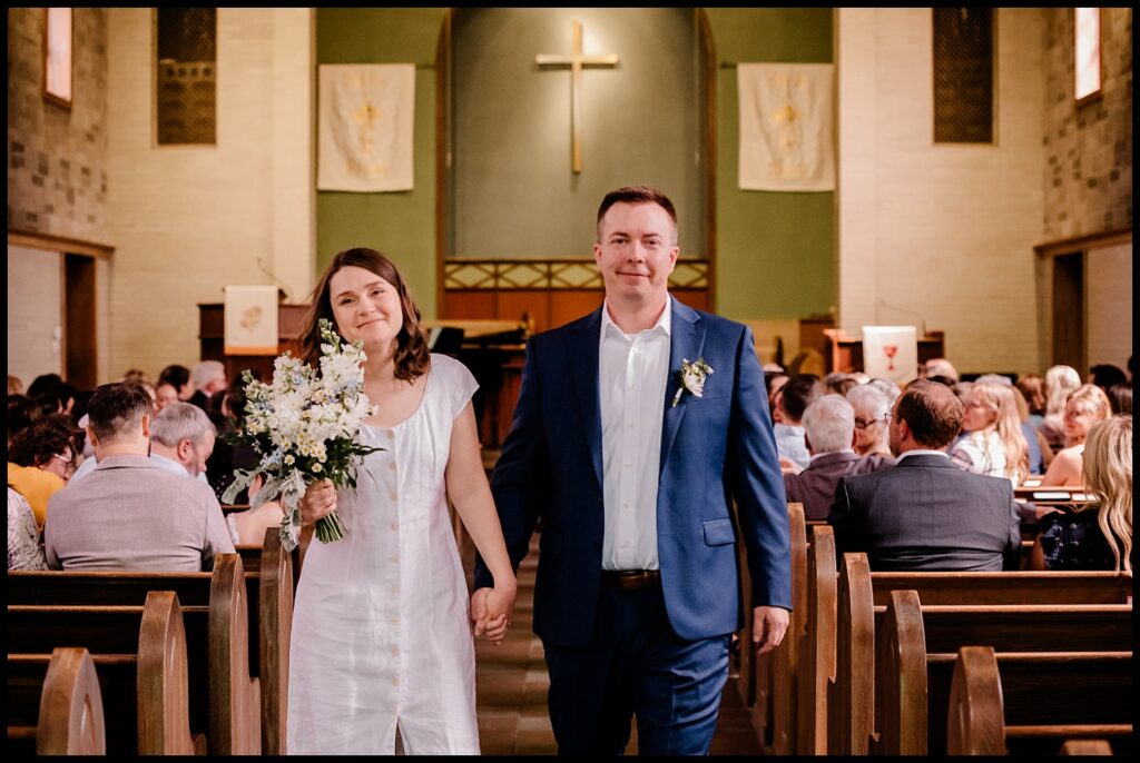 Jess and Chris, holding hands, walk down the aisle of a church filled with seated guests. Jess holds a bouquet and wears a white dress, while Chris wears a blue suit. A large cross hangs at the front of the church.