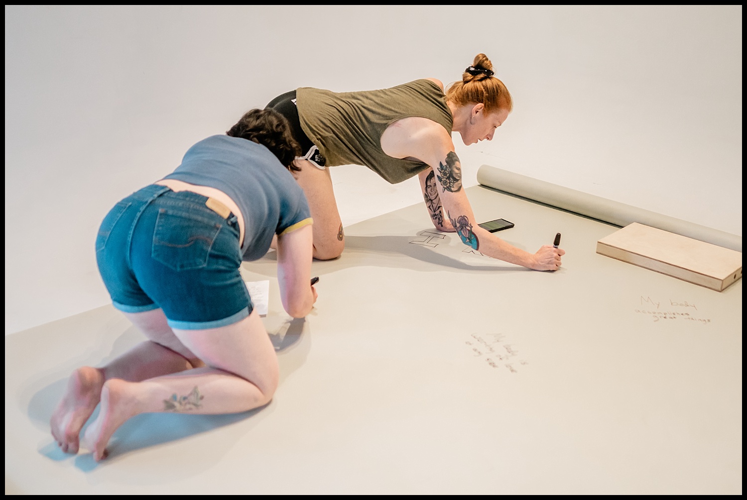 Two people kneel on the floor, writing or drawing on a large white sheet of paper with markers—capturing a scene of creativity and body acceptance. A phone and some papers are beside them, adding to this candid Nashville Boudoir-inspired moment.