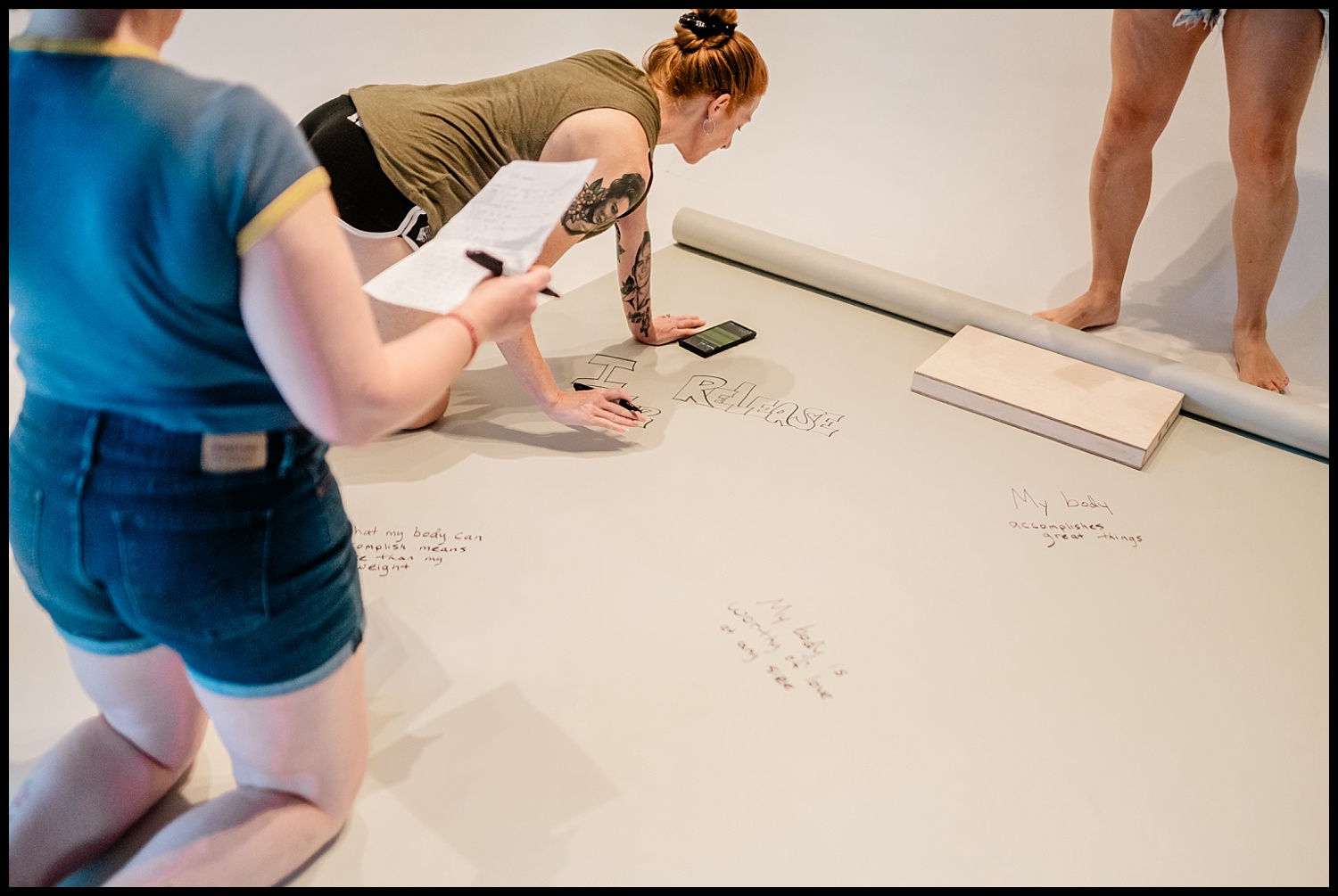 Two people kneel on the floor, writing messages about body acceptance on a large white sheet. One holds a notepad, another uses a phone. Legs of a third are visible nearby. Messages and drawings cover the sheet in this Nashville Boudoir empowerment shoot.