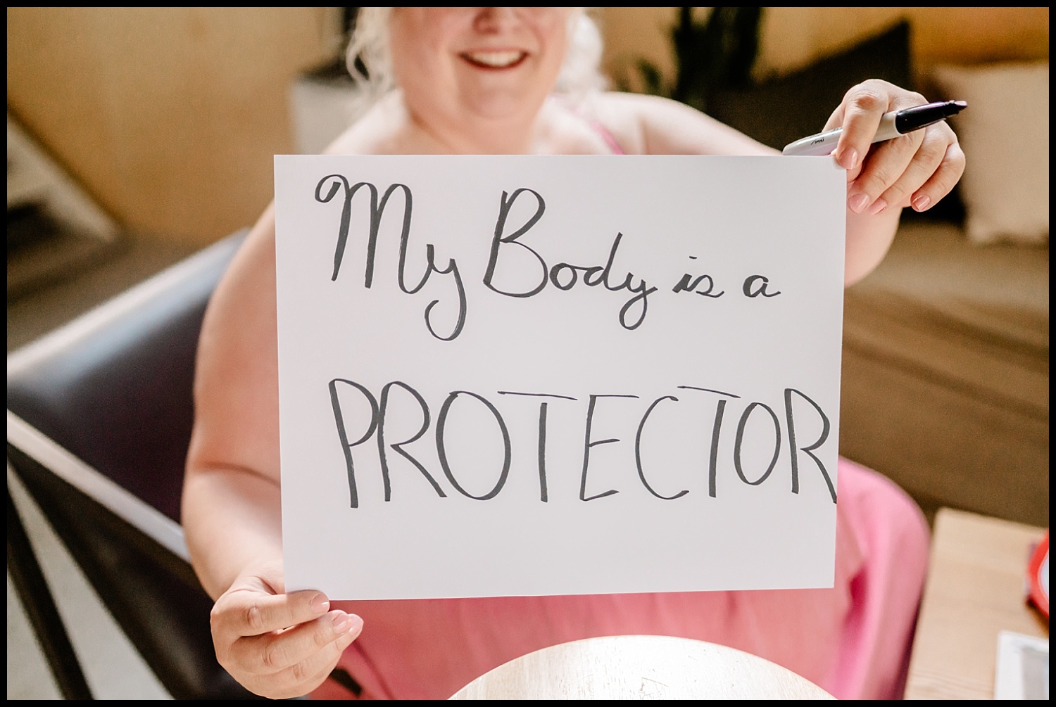 A smiling person in a pink dress holds a sign that reads, My Body is a PROTECTOR, celebrating self-love and body acceptance with bold letters.