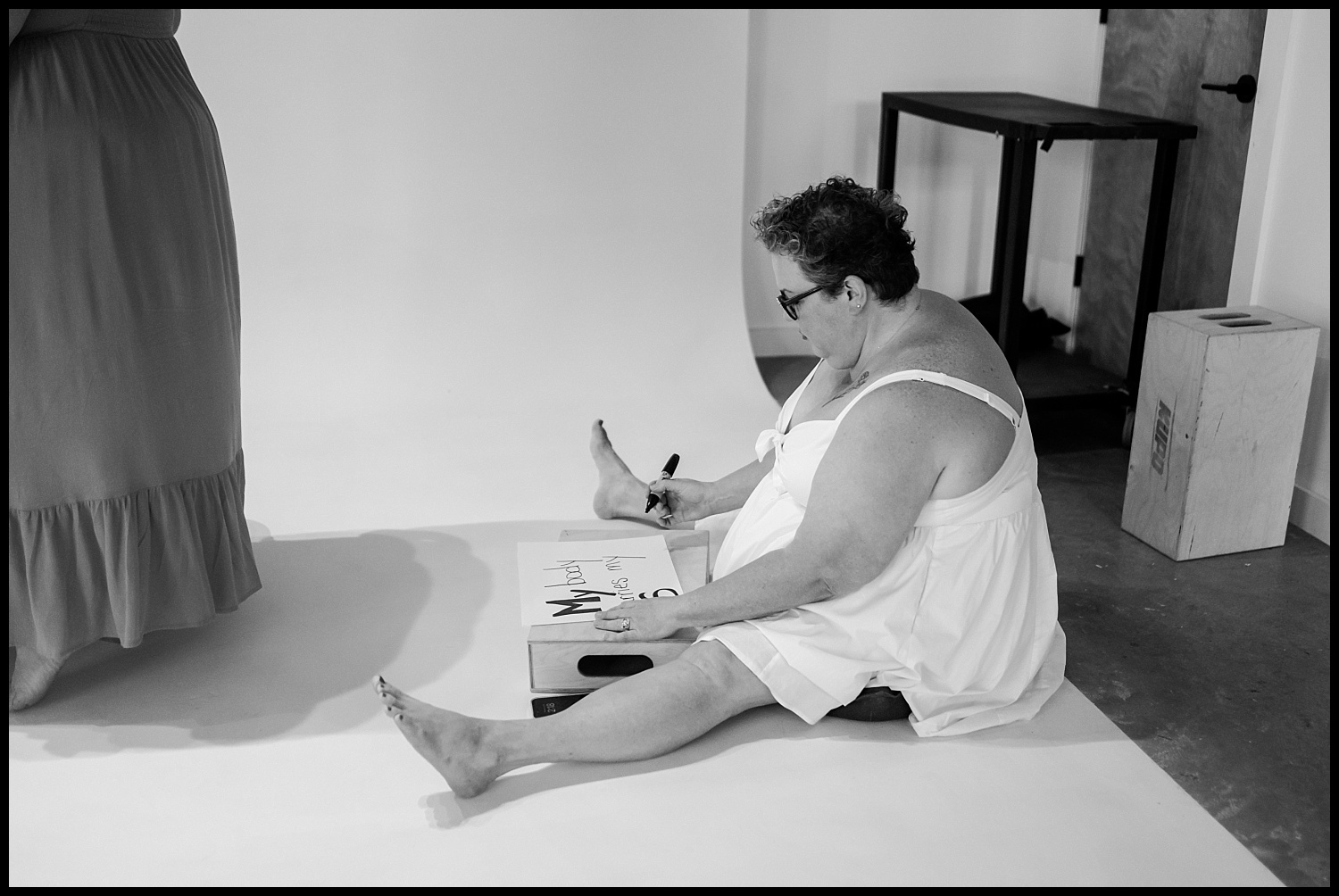 A woman in a white dress sits on the floor with her legs outstretched, writing on a poster with a marker. Another person in a long dress stands nearby. The minimalist indoor studio hints at an empowerment shoot or Nashville boudoir session.
