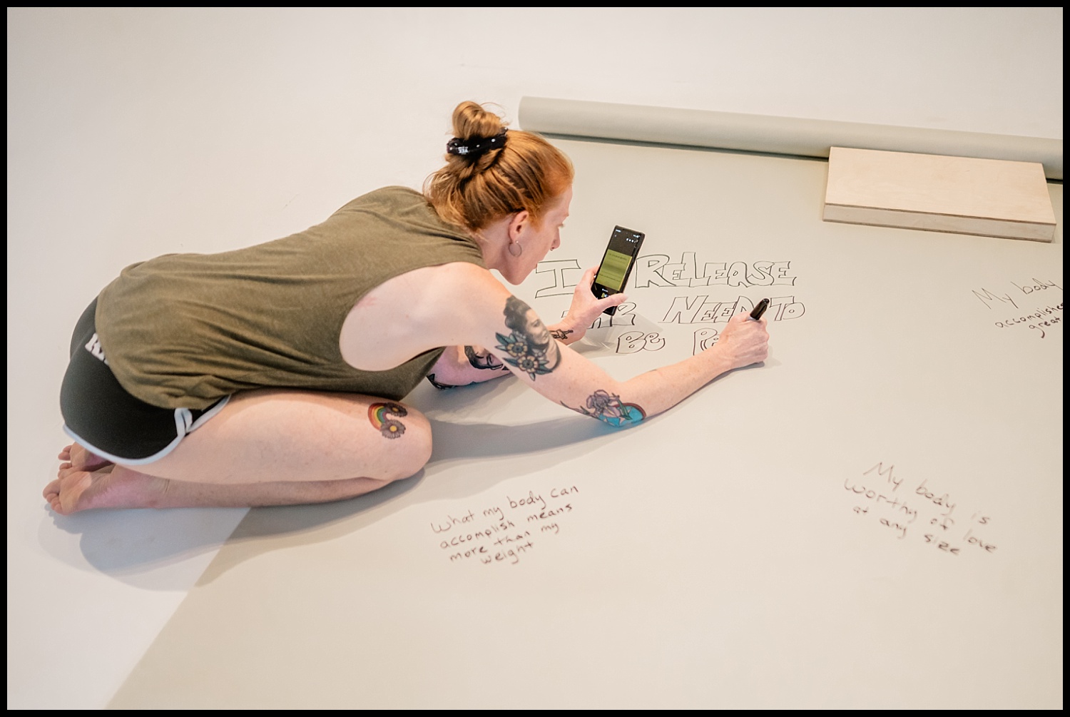 A person with tattoos kneels on the floor, writing messages of self-love and drawing on a large sheet of paper. They hold a smartphone and wear a green tank top and black shorts. Handwritten notes surround them.