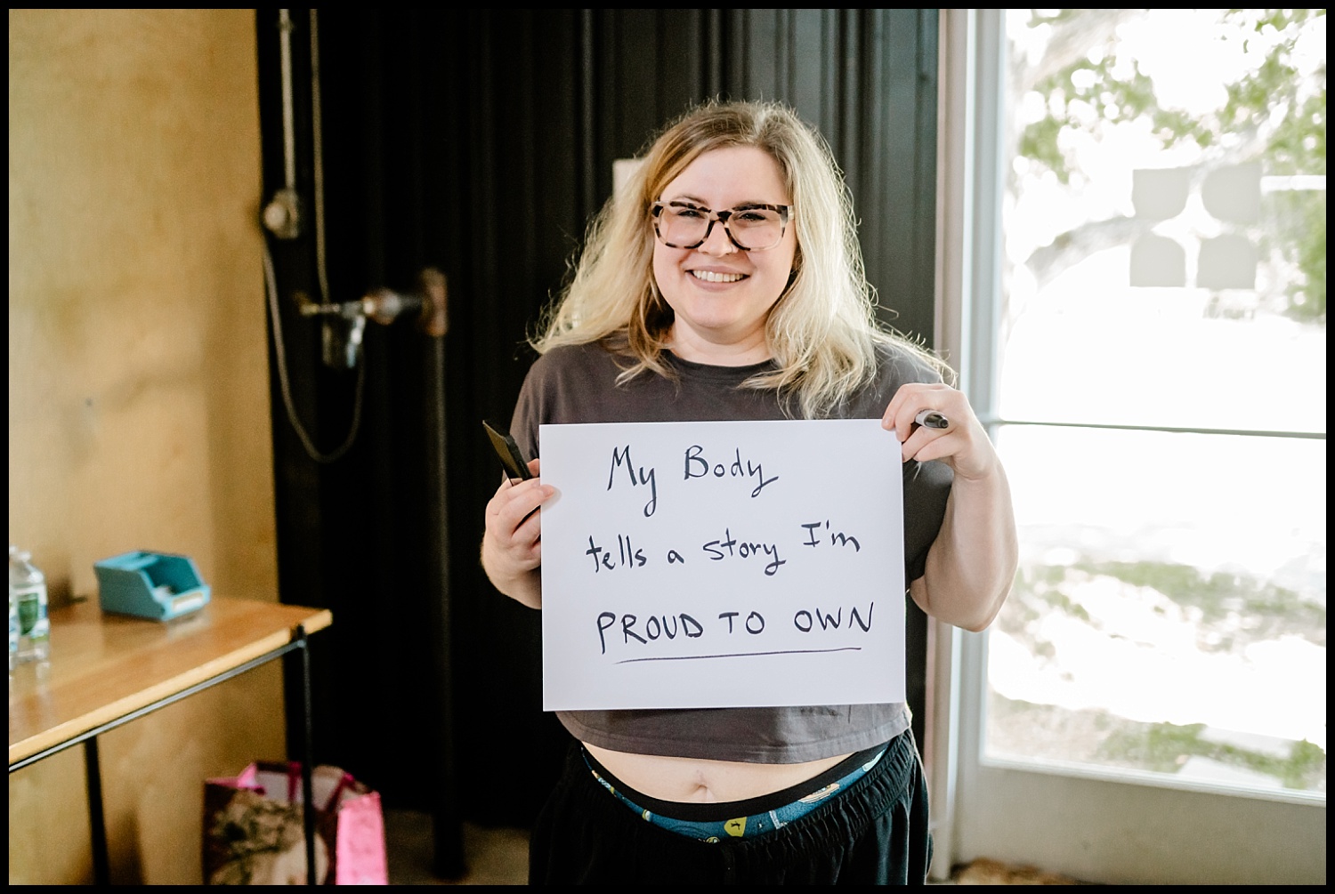 A smiling person with glasses and blond hair holds a sign that says, My body tells a story Im proud to own during an Empowerment Shoot, standing indoors near a window with trees visible outside.