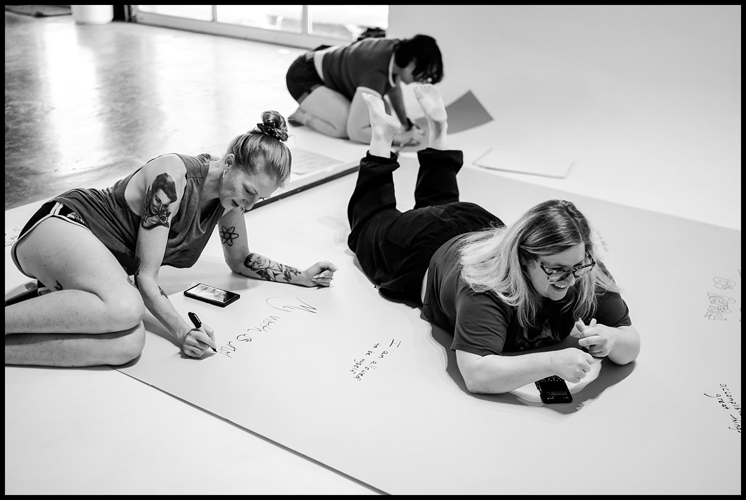 Three people are on the floor writing on large sheets of paper. One woman with a tattoo writes with a marker, embracing self-love, while another lies on their stomach writing and a third sits in the background, also writing.