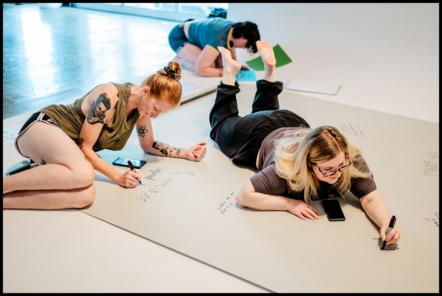 Three people lie on the floor writing on large sheets of paper in an Empowerment Shoot. Two are in the foreground, one with tattoos, the other with long blonde hair and glasses. A third person writes on a separate sheet in the background.