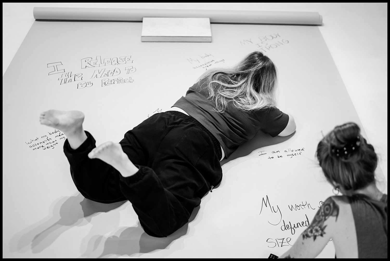 A woman lies on her stomach on a large sheet of paper, writing messages with a marker in this Nashville Boudoir empowerment shoot. Handwritten notes and affirmations reflecting body acceptance are visible nearby. The image is in black and white.