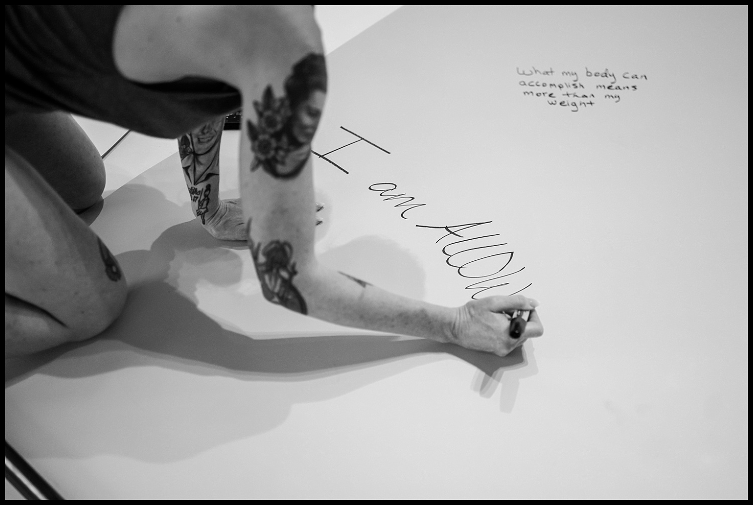 A person with tattoos writes I am ALLOW on a white surface during an empowerment shoot. Nearby, a message reads, What my body can accomplish means more than my weight. The black and white scene radiates self-love.