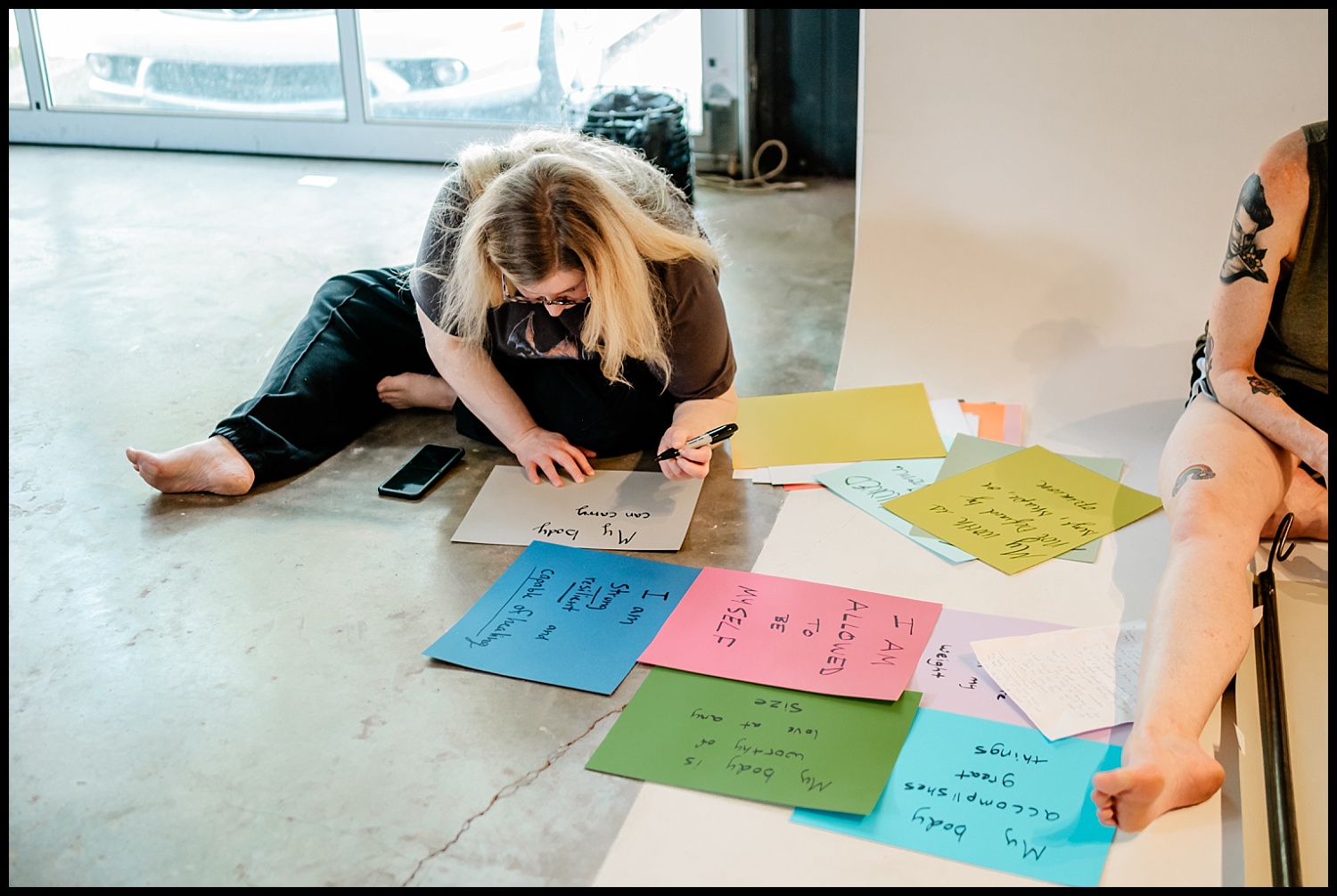 A person sits on the floor, writing on a large piece of paper surrounded by colorful sheets filled with self-love notes. Another person with a tattooed arm is partially visible. A phone and window are nearby, capturing an empowerment shoot moment.