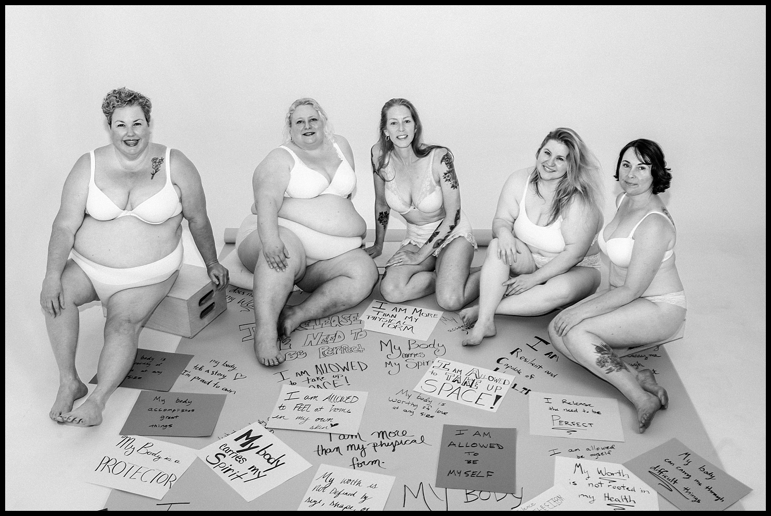 A black and white photo of five women in underwear sitting together, smiling. Surrounded by handwritten signs with empowering self-love messages on the floor, this Nashville boudoir scene radiates body acceptance.