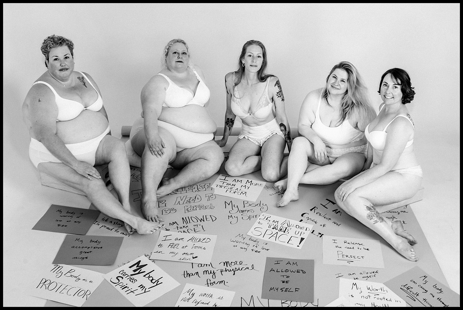 Five women in white underwear sit together, smiling and posing confidently for an empowerment shoot. Around them are handwritten signs with body-positive affirmations such as “My body is worthy.” Black and white photo.